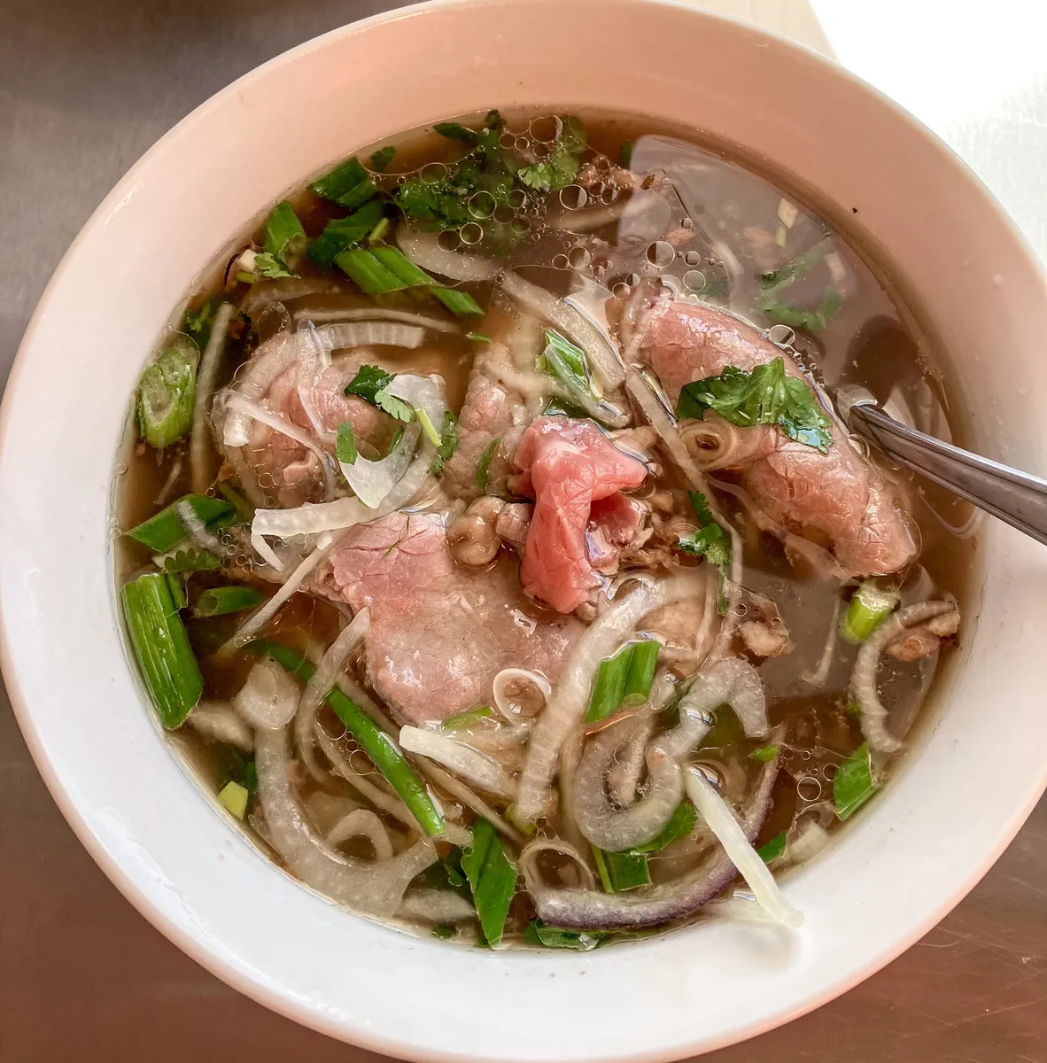 A bowl of beef phở with sliced beef, rice noodles, onions, scallions, and fresh herbs in clear broth