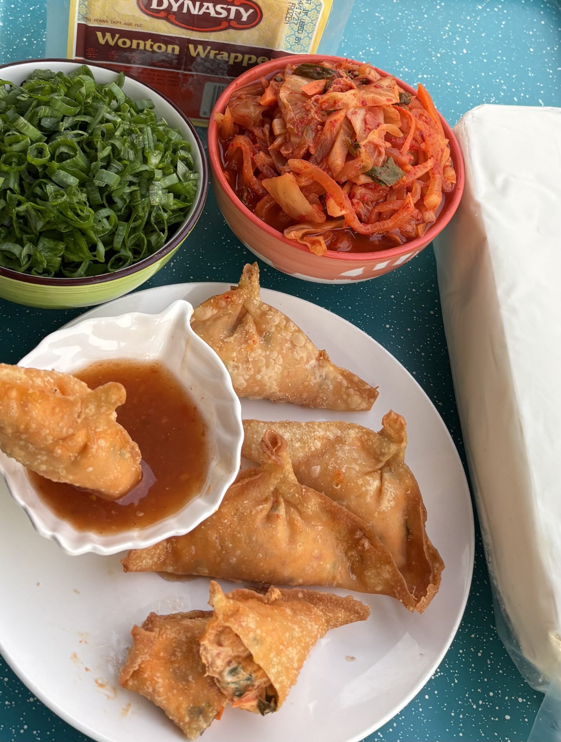 Crispy golden Hungry Lotus kimchi wontons on a white plate with sweet chili dipping sauce, beside bowls of chopped green onion and kimchi, a package of wonton wrappers, and a block of cream cheese